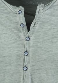 Maglietta henley grigia chiara con scollatura a girocollo, caratterizzata da cinque bottoni blu, una texture morbida e sottili righe verticali.