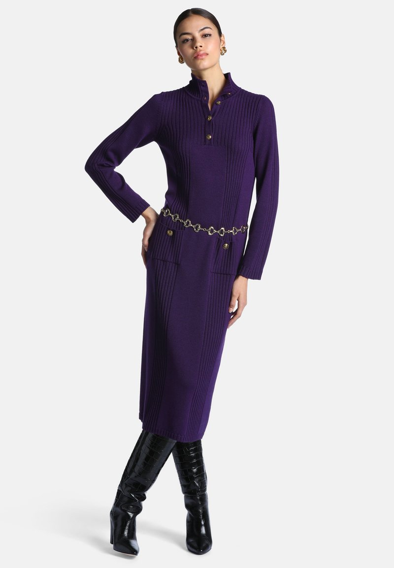 Femme portant une robe moulante en maille côtelée violette avec des boutons dorés et une ceinture chaîne, associée à des bottes en cuir noires montantes jusqu'au genou, debout avec une main sur la hanche.