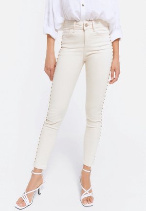 Femme portant un jean skinny taille haute blanc cassé avec des clous métalliques le long des coutures extérieures, associé à des sandales à talons blanches à brides.