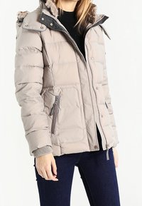 Mujer vistiendo una chaqueta de invierno acolchada beige con capucha forrada de piel, bolsillos con cremallera, suéter negro y jeans azul oscuro, de pie contra un fondo blanco.