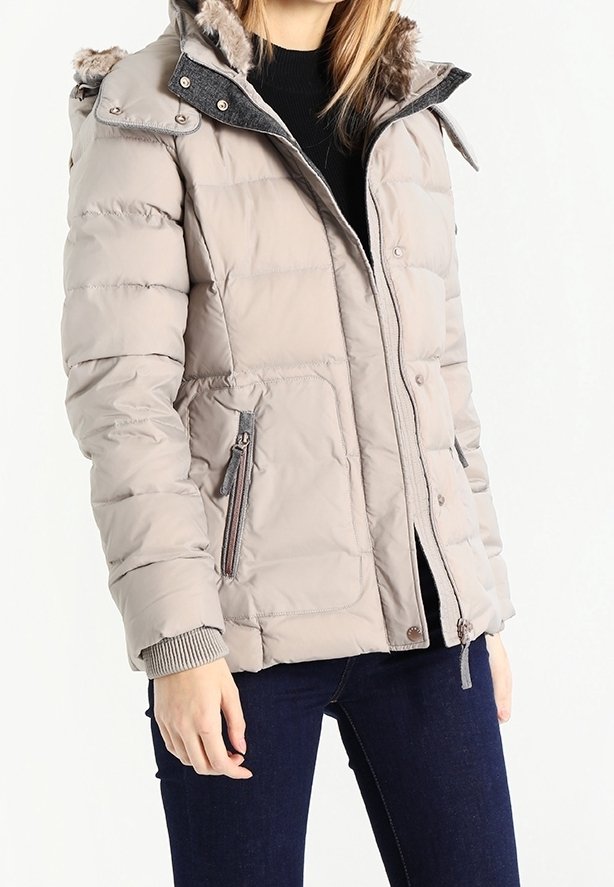 Mujer vistiendo una chaqueta de invierno acolchada beige con capucha forrada de piel, bolsillos con cremallera, suéter negro y jeans azul oscuro, de pie contra un fondo blanco.