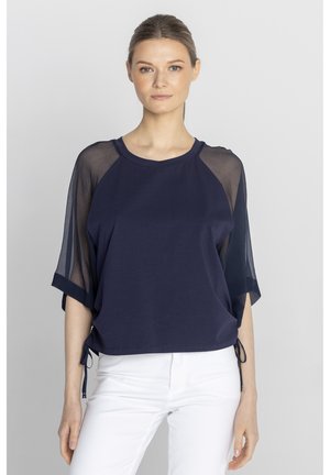 Kobieta w granatowej bluzce z przejrzystymi rękawami i białych spodniach, stojąca na jednolitym jasnoszarym tle.