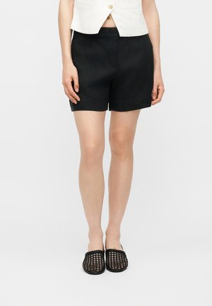 Kvinde iført sorte skræddersyede shorts, hvid cropped vest og sorte vævede slip-ons, stående mod en lys baggrund.