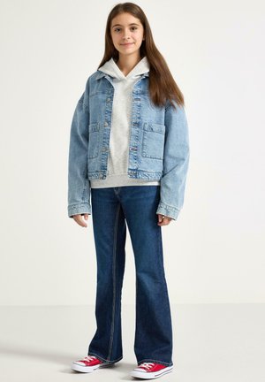 Lyseblå denimjakke over en grå hættetrøje, kombineret med mørkeblå flare-jeans og røde sneakers, med synlige sømme og lommer.