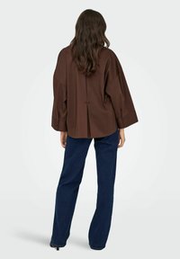 Femme aux longs cheveux bruns vue de dos, portant un chemisier marron ample à manches larges et un jean bleu foncé à jambes droites.