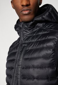 Črn puffer telovnik z vodoravno prešito teksturo, visokim ovratnikom in sprednjim zadrgo. Gladek, sijoč material z lahkotnim videzom.