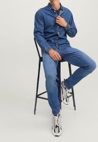 Modrá džínová košile a džíny s límečkem na knoflíky, doplněné vícebarevnými teniskami, sedící na kovové stoličce s opotřebovaným povrchem.