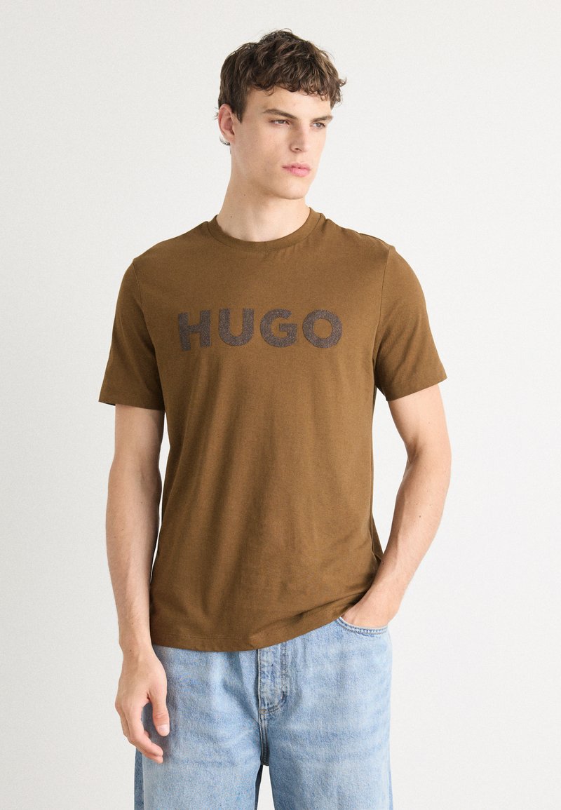 Barna, rövid ujjú póló, merész fekete "HUGO" felirattal; puha anyagból készült, kerek nyakkivágással és laza szabással.