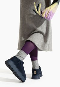 Granatowe zamszowe buty na grubej, ciemnej gumowej podeszwie, noszone z szarymi prążkowanymi skarpetkami i fioletowymi rajstopami pod szarą sukienką.