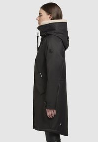 Schwarze lange Parka-Jacke mit einer mit Fell gefütterten Kapuze, strukturiertem Stoff, Seitentaschen und einem Markenpatch am Ärmel. Elegantes Design, mit Knöpfen an den Ärmeln.