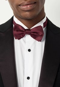 Noeud papillon en satin bordeaux porté sur une chemise blanche avec des boutons noirs ; associé à une veste de smoking noire, mettant en avant les détails de la tenue formelle.
