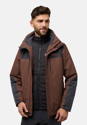 Braune wasserdichte Jacke mit grauen Akzenten, abnehmbarer Kapuze und Reißverschlüssen. Getragen über einer schwarzen isolierten Weste mit gestepptem Muster.