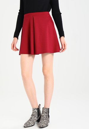 Mujer con minifalda plisada roja, top de manga larga negro y botines de estampado de leopardo, de pie contra un fondo blanco.