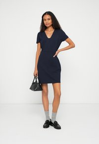 Tamsiai mėlyna aptempianti suknelė su V formos iškirpte ir trumpomis rankovėmis. Derinama su pilkomis kojinėmis ir juodais loaferiais. Laiko mažą juodą rankinę.