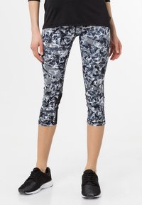 Leggings capri noir et blanc avec un motif abstrait audacieux de feuilles et de rayures, fabriqués en tissu extensible. Associés à des chaussures de sport noires.