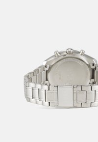 Srebrna ura iz nerjavečega jekla z brušenim vzorcem, povezavnim trakom in okroglo ohišjem. Ima tri gumbke in gravirano oznako "OTPORNO NA VODO" na hrbtni strani.