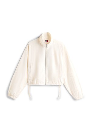 Veste légère blanche avec fermeture éclair frontale, poignets élastiques, ourlet à cordon de serrage, et petit logo rouge, blanc et bleu sur le côté gauche de la poitrine.