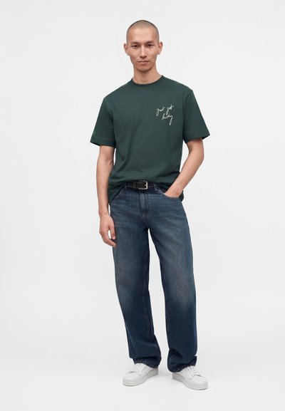 Homme à la tête rasée portant un t-shirt vert foncé avec un texte blanc "just get lucky,", jean bleu, ceinture noire et baskets blanches, debout avec une main dans la poche.