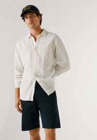 Jeune homme portant une chemise blanche à manches longues boutonnée, un short foncé et une casquette de baseball noire, debout avec une main derrière le dos.