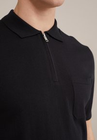 Black knit polo shirt with a zip collar, featuring a subtle pocket on the left chest and a soft texture. Simple, modern design.