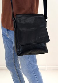 Borsa a tracolla in pelle nera con chiusura a patta, texture liscia e attacco per tracolla in argento. Presenta un branding sottile.