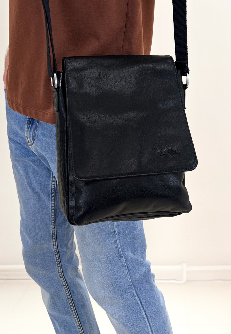 Borsa a tracolla in pelle nera con chiusura a patta, texture liscia e attacco per tracolla in argento. Presenta un branding sottile.