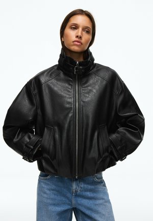 Femme portant une veste en cuir noire avec un col montant et une fermeture éclair, les mains dans les poches, associée à un jean bleu sur un fond uni.