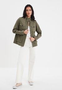 Giacca utility verde oliva con colletto alto, due tasche sul petto e dettagli in bottone, indossata sopra una camicia bianca e abbinata a pantaloni ampi color crema.