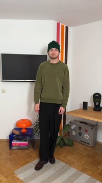 Maglione verde oliva a trama grossa, pantaloni neri ampi e berretto a righe verde. Scarpe marroni. In piedi in una stanza con arredamento moderno.