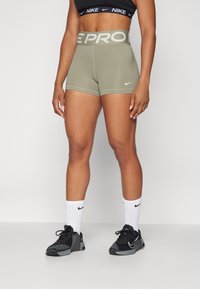Olivově zelené šortky Nike Pro s širokým elastickým pasem, na kterém je v světle šedé barvě nápis 'PRO'. Doplňují je bílé ponožky Nike a černé sportovní boty.