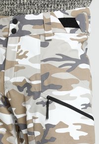Beige och vita byxor med kamouflage-mönster, en dragkedjeficka, två svarta knappar i midjan och en texturerad midja för stabilitet.
