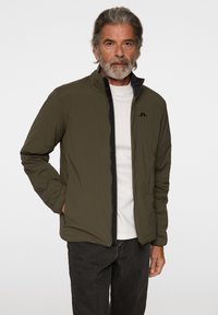 Olivgrön lätt jacka med dragkedja framtill och ståkrage, med en svart accent längs dragkedjan och en logotyp på bröstet.