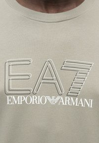 Közelkép egy bézs pulóverről, amelyen fehér kontúrral rajzolt "EA7" és "EMPORIO ARMANI" feliratok, valamint egy kis saslogó látható középen.