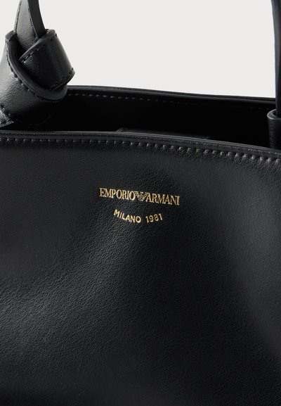 Μαύρη δερμάτινη τσάντα με ραμμένα άκρα, διακοσμημένη με χρυσό λογότυπο Emporio Armani και το κείμενο "Milano 1981" κοντά στο επάνω κέντρο.