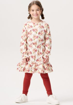 Robe florale à manches longues, avec un ourlet volanté et une taille élastique ; imprimée de motifs roses et verts sur un fond crème. Associée à des leggings rouges et des baskets blanches.