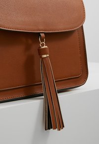 Brun handväska i konstläder med klaffstängning, som har en guldfärgad metallaccent och en lång brun tofsdetalj. Texturerad yta.