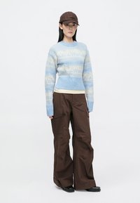 Šviesiai mėlynas megztas svarkelis su susiūta juosta, rudi krovinių kelnės ir ruda kepurė, su logotipo raštu per svarkelio priekį.