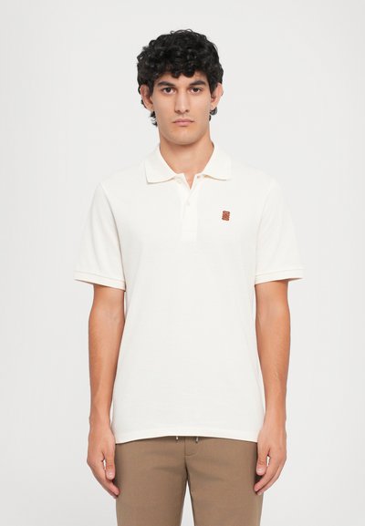 Light beige polo shirt made from soft cotton, featuring a collar and short sleeves. Small brown logo embroidered on the chest.