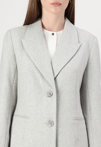 Helehall herringbone särk, millel on nööbiga kaelus, kahe nööbiga kinniti ja esikotid, valmistatud tekstureeritud kangast.