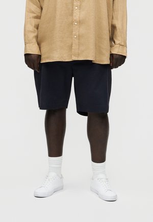 Άτομο που φοράει μπεζ πουκάμισο με μακριά μανίκια, σκούρα μπλε σορτς, λευκές κάλτσες και λευκά αθλητικά παπούτσια, στέκεται μπροστά σε απλό φωτεινό φόντο.