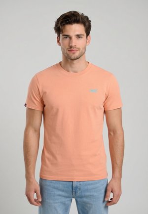 Giovane uomo con capelli corti e mossi indossa una maglietta a maniche corte color pesca e jeans azzurri chiari su uno sfondo grigio uniforme.