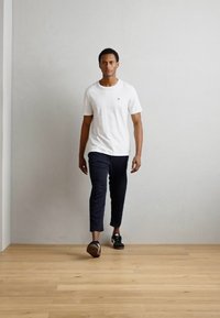 Witte t-shirt met korte mouwen en een klein logo, gecombineerd met donkere slim-fit broek. Model draagt zwarte sneakers en staat op een lichte houten vloer.