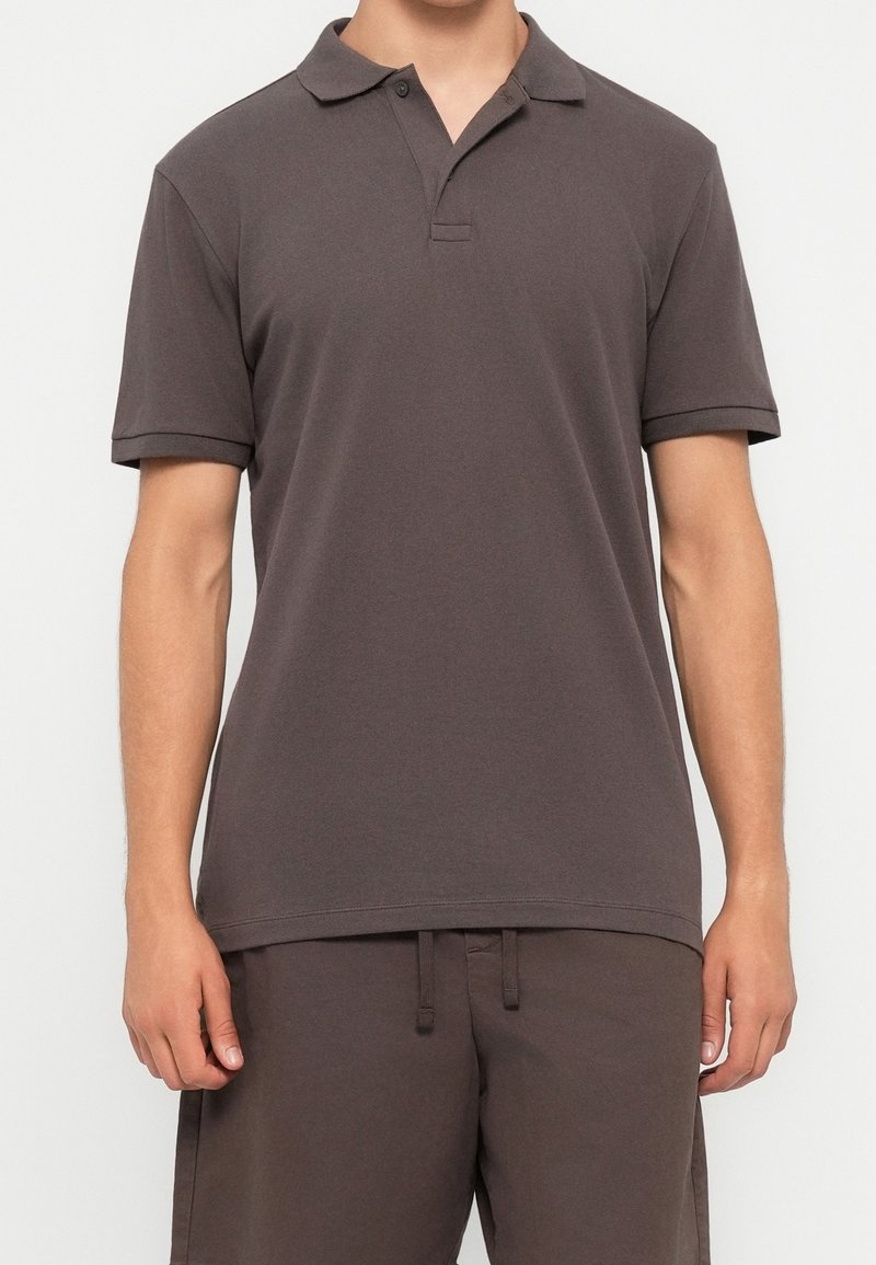 Homme portant un polo marron foncé uni à manches courtes et un pantalon à cordon assorti, debout devant un fond blanc.