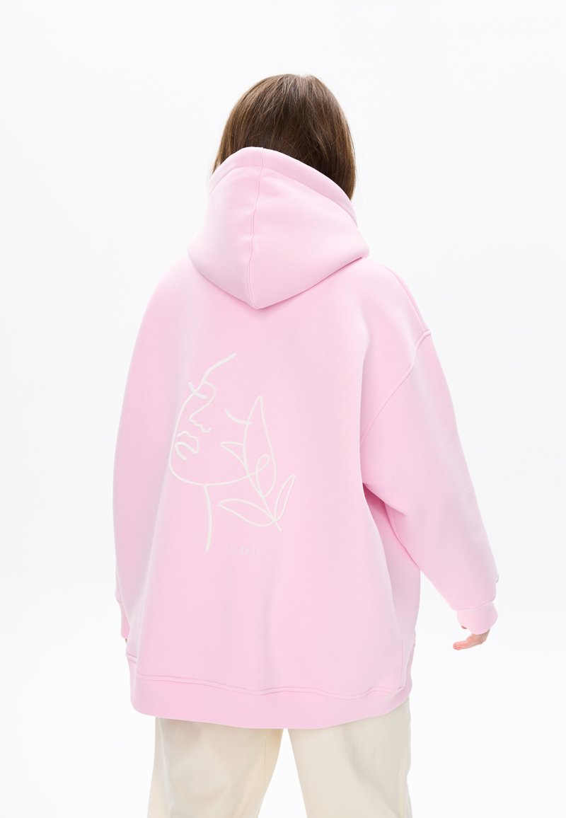 Oversized roze hoodie met een trekkoord in de capuchon, met een witte lijntekening van een gezicht en bladeren op de achterkant. Zachte textuur.