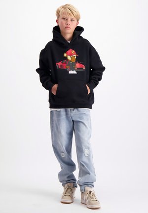 Zwarte hoodie met een afbeelding van een cartoonbeer in een rode pet naast een rode sportwagen, gecombineerd met lichtblauwe distressed jeans en beige sneakers.