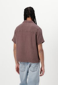Kortærmet skjorte i struktureret brun stof med en afslappet pasform, der har krave og lige kant. Båret sammen med lyseblå denimjeans.