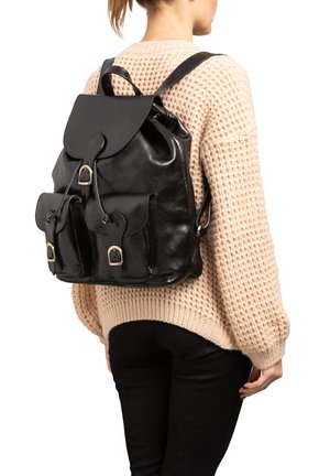 Vrouw draagt een beige gebreide trui en zwarte broek, en heeft een zwarte leren rugzak met voorvakken met gespen bij zich.