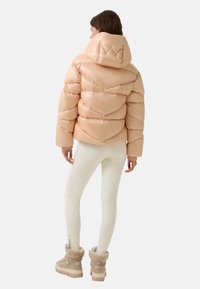 Różowa kurtka puchowa z wysokim kołnierzem, pikowanym wzorem i ściągaczami, w zestawieniu z kremowymi legginsami i beżowymi butami ocieplanymi futrem.
