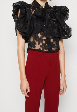 Schwarze transparente Bluse mit großen Blumenmustern und gepufften Ärmeln, kombiniert mit hoch taillierten roten Hosen.