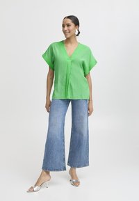 Top verde a collo a V con texture e maniche corte, abbinato a jeans in denim blu a gamba larga e sandali con tacco argentati, su uno sfondo neutro.